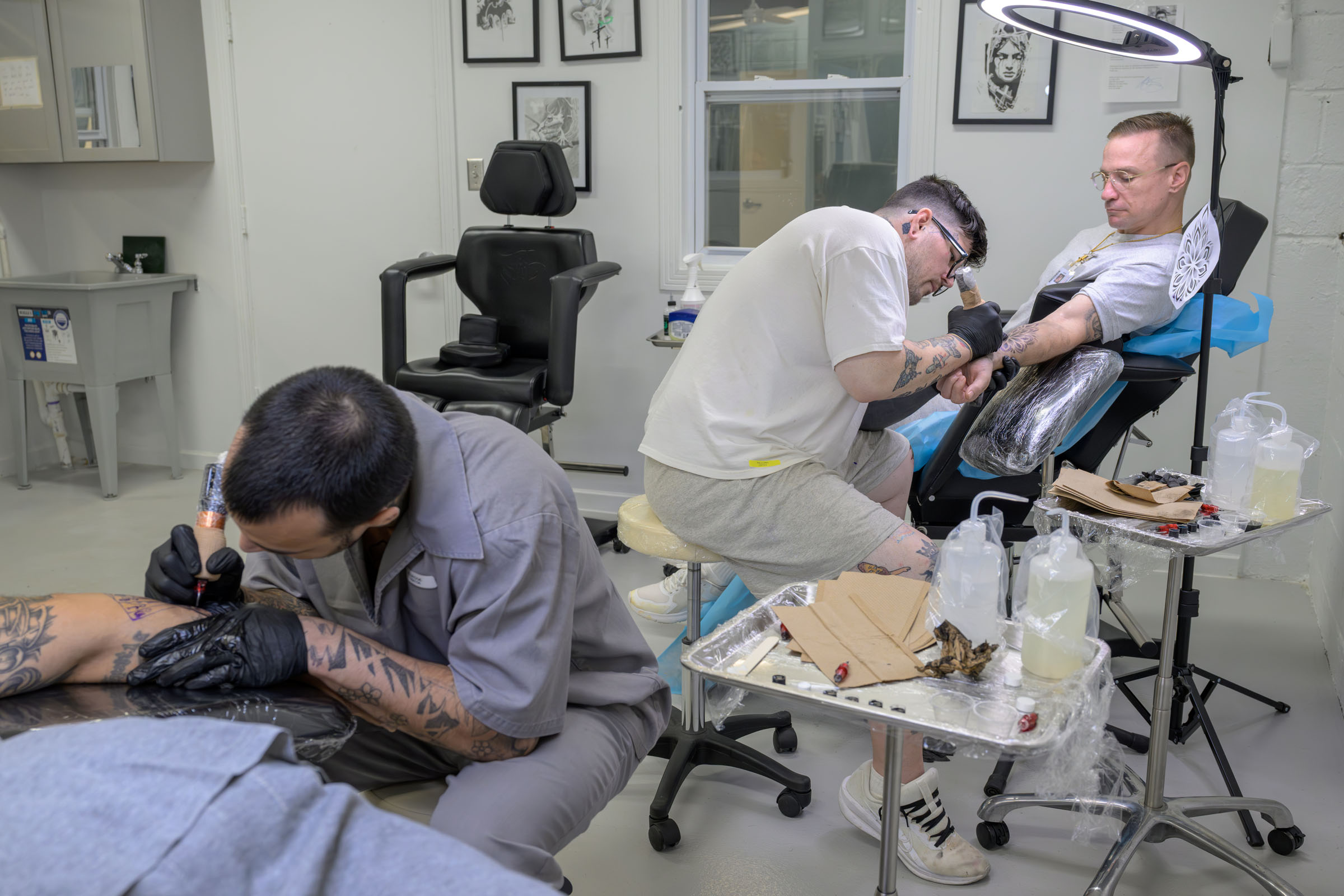 Two tattoo artists work on two clients in a tattoo studio.
